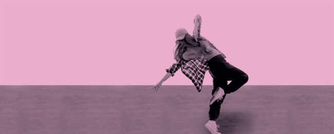 Ally Pally Performing Arts School. A dancer in casual attire strikes a dynamic pose against a solid pink backdrop. She wears a cap, a plaid shirt, and sneakers, conveying energy and movement.
