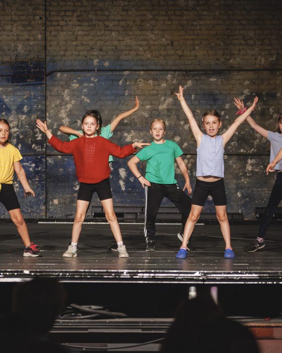 Seven children energetically dance on stage in colorful outfits against a textured brick wall, conveying excitement and joy in a lively performance.