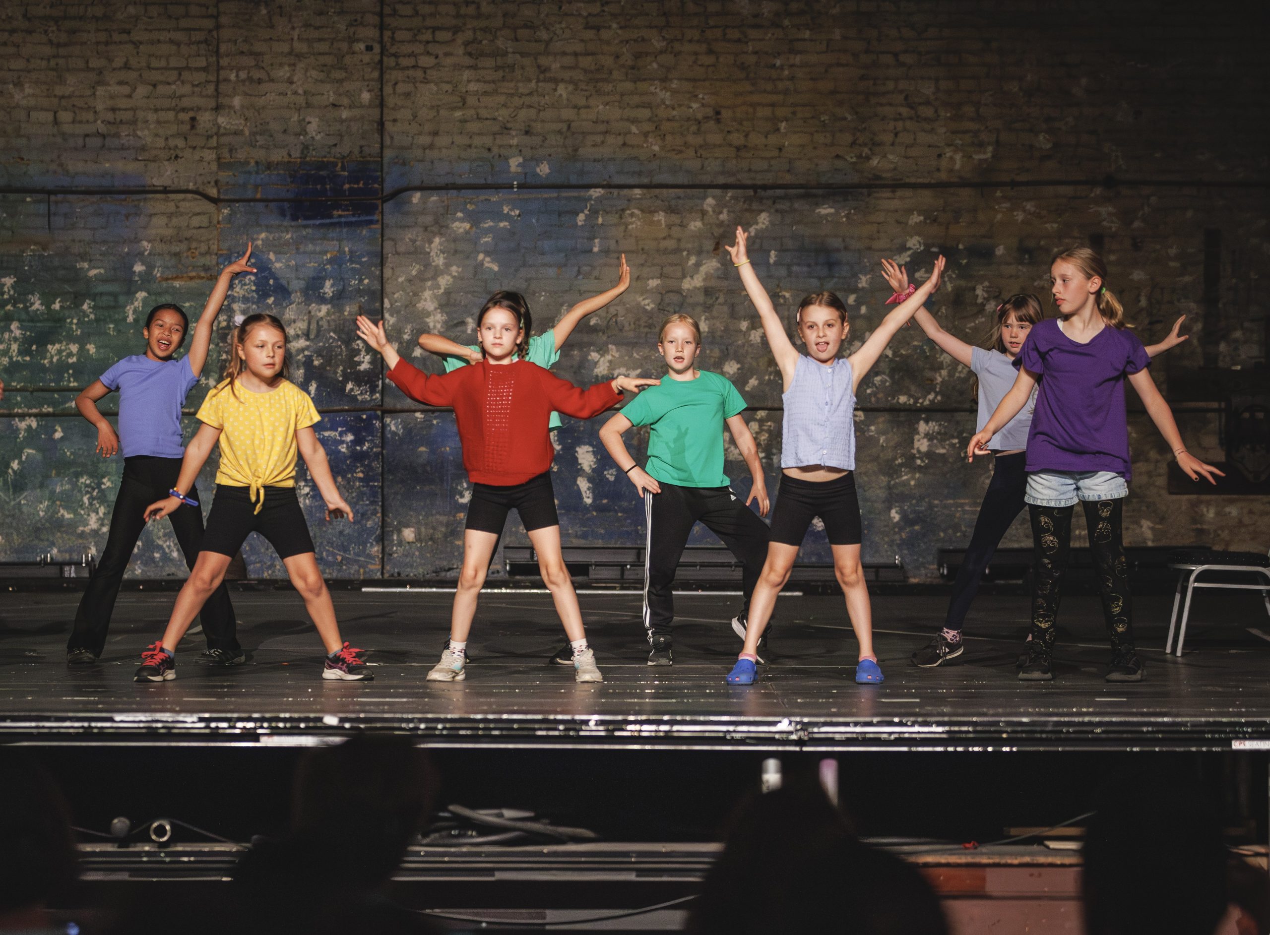 Seven children energetically dance on stage in colorful outfits against a textured brick wall, conveying excitement and joy in a lively performance.