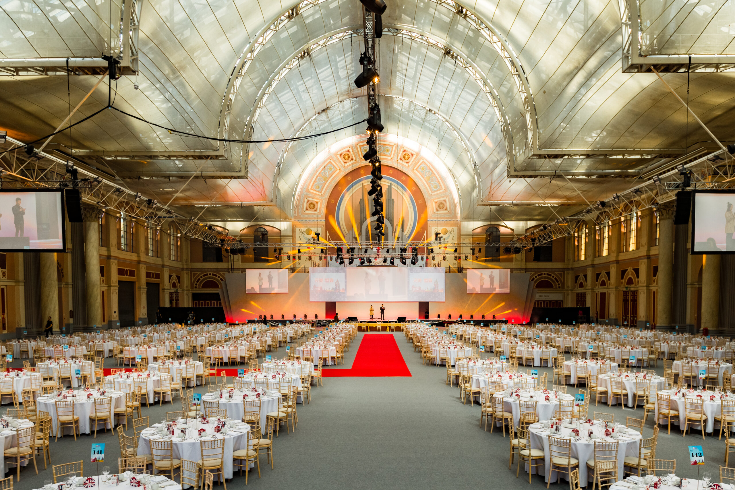 Large hall with columns ready for a formal event with round tables, chairs, stage, and a red carpet.