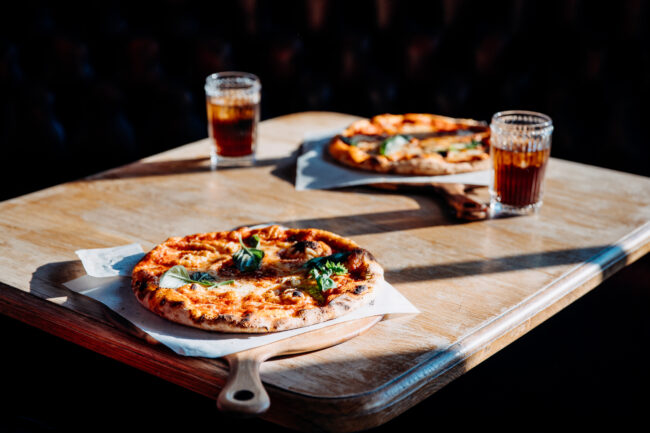 Two pizzas on a table with two glasses of fizzy drink in the sun.