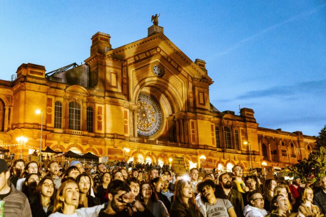 A large group of people in front of a Alexandra Palace singing at sunset.