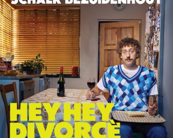 Man with curly hair and mustache in a blue patterned sweater vest, sits at a kitchen table holding a wine glass. A bottle and houseplants are in the background.