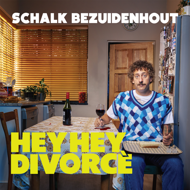 Man with curly hair and mustache in a blue patterned sweater vest, sits at a kitchen table holding a wine glass. A bottle and houseplants are in the background.