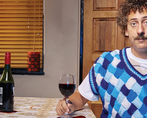 Man with curly hair and mustache in a blue patterned sweater vest, sits at a kitchen table holding a wine glass. A bottle and houseplants are in the background.