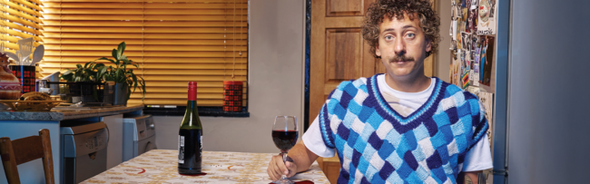 Man with curly hair and mustache in a blue patterned sweater vest, sits at a kitchen table holding a wine glass. A bottle and houseplants are in the background.