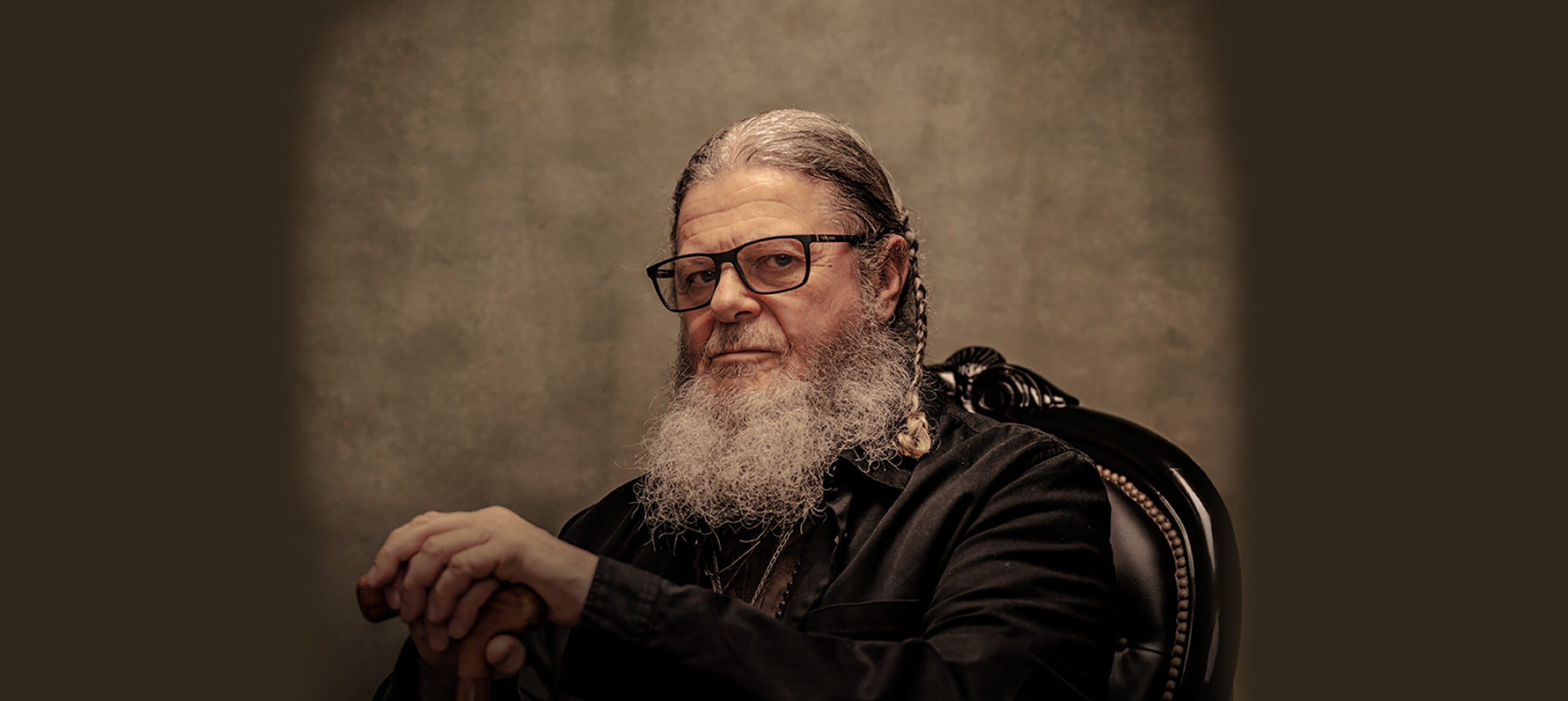 A portrait of Argentinian composer Gustavo Santaolalla. He has a long grey beard and glasses sits on a black ornate chair, wearing dark clothing and holding a wooden cane against a neutral background.