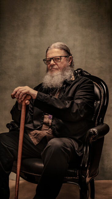 A portrait of Argentinian composer Gustavo Santaolalla. He has a long grey beard and glasses sits on a black ornate chair, wearing dark clothing and holding a wooden cane against a neutral background.