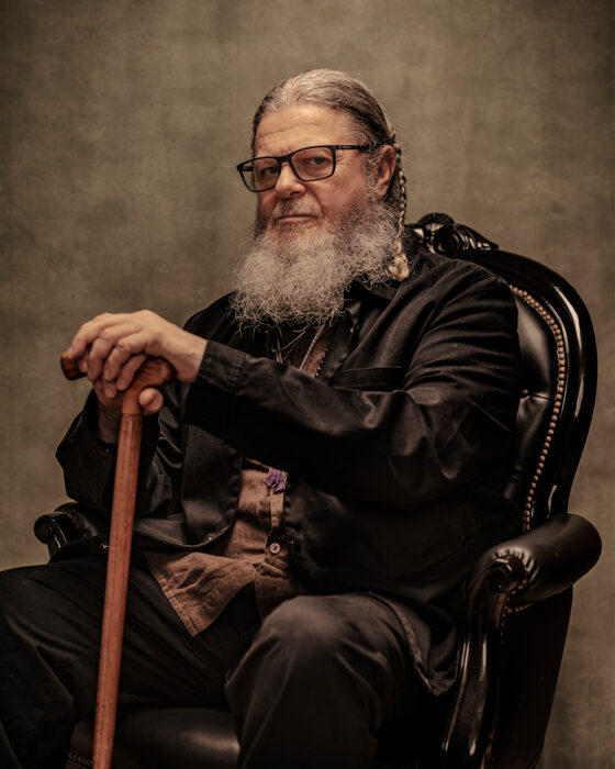 A portrait of Argentinian composer Gustavo Santaolalla. He has a long grey beard and glasses sits on a black ornate chair, wearing dark clothing and holding a wooden cane against a neutral background.
