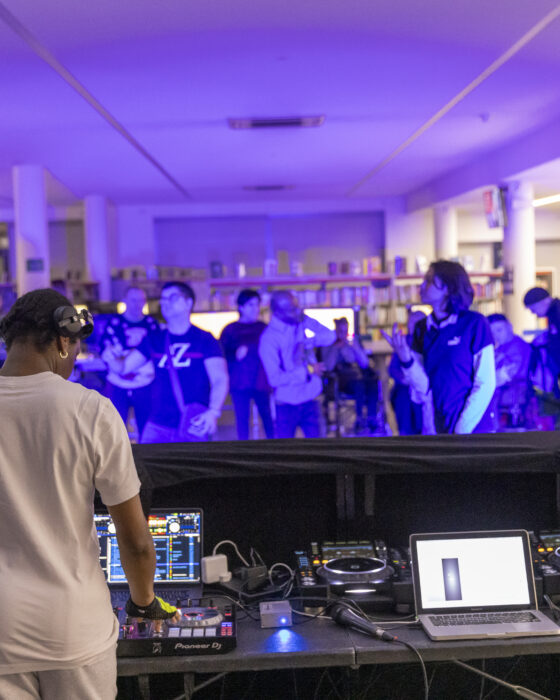 DJ in white stands at a console with turntables and a laptop, performing in a purple-lit room. A diverse crowd enjoys the music at the back.
