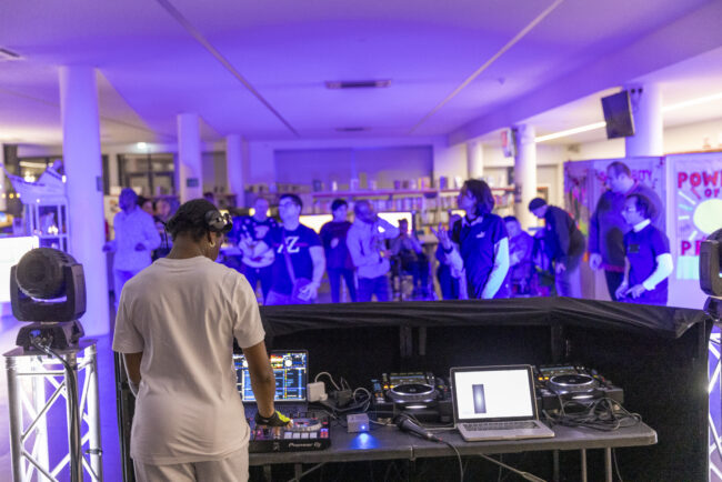 DJ in white stands at a console with turntables and a laptop, performing in a purple-lit room. A diverse crowd enjoys the music at the back.