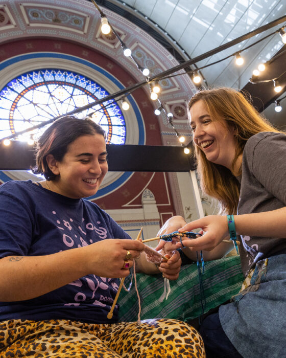 Two people happily knitting together in a bright hall, decorated with string lights and a stained glass window, conveying a joyful, creative atmosphere.