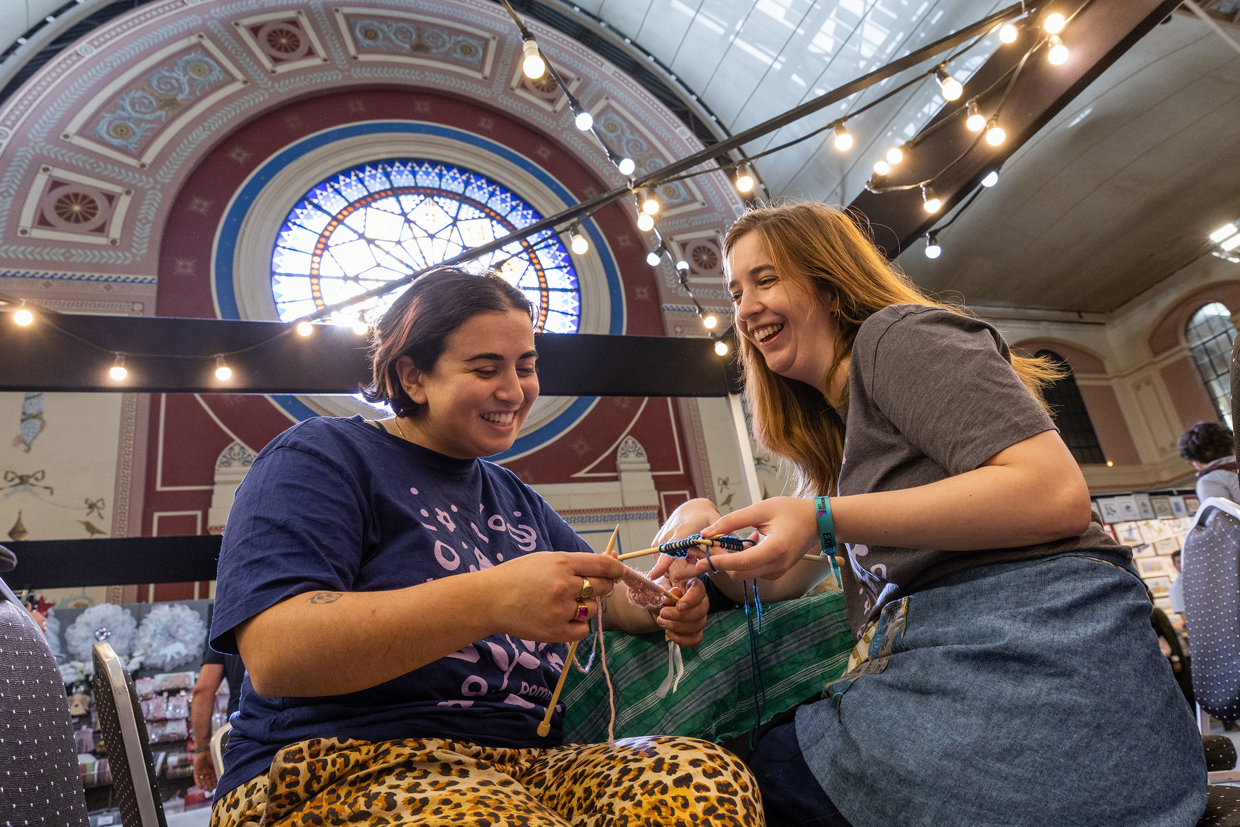 Two people happily knitting together in a bright hall, decorated with string lights and a stained glass window, conveying a joyful, creative atmosphere.