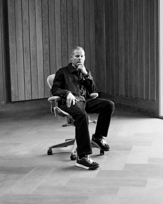 A person sits pensively in a minimalist room with wooden walls, beside a large window overlooking a grassy landscape, conveying calm introspection.