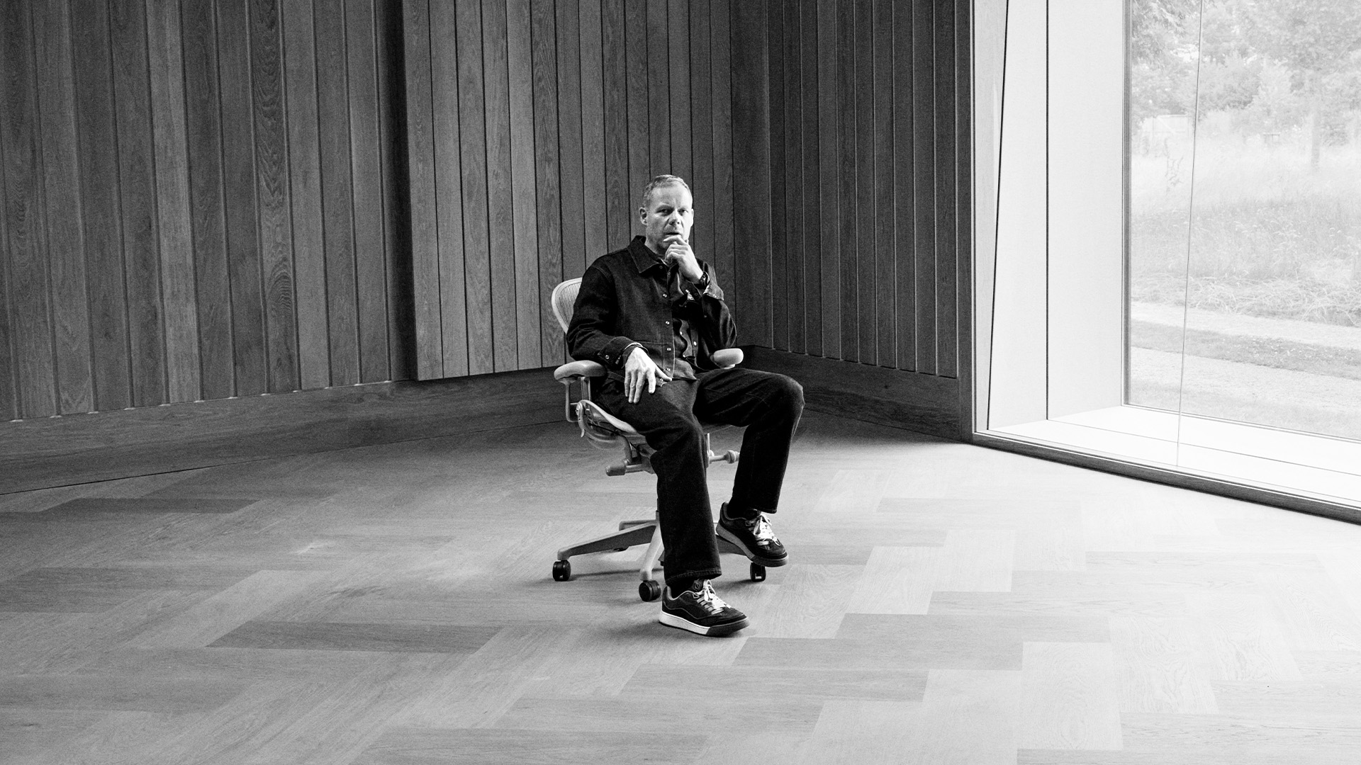 A person sits pensively in a minimalist room with wooden walls, beside a large window overlooking a grassy landscape, conveying calm introspection.