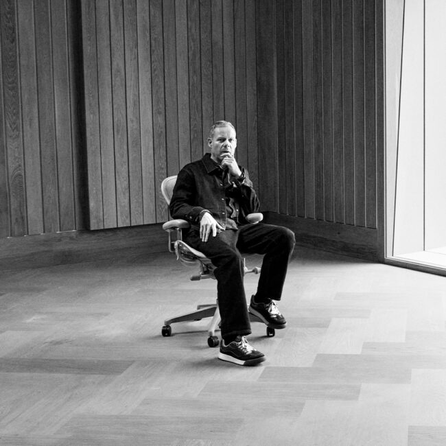 A person sits pensively in a minimalist room with wooden walls, beside a large window overlooking a grassy landscape, conveying calm introspection.