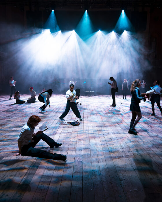 Dimly lit theater with wooden floors and a high ceiling. People are scattered, engaged in various activities. Spotlights illuminate the scene, creating a dramatic atmosphere.