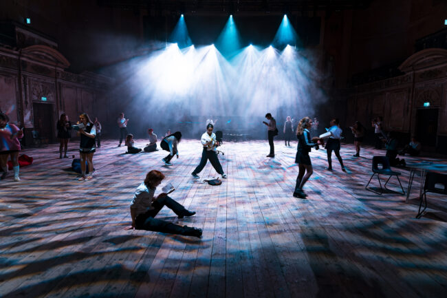 Dimly lit theater with wooden floors and a high ceiling. People are scattered, engaged in various activities. Spotlights illuminate the scene, creating a dramatic atmosphere.
