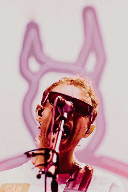 The lead singer from band Wunderhorse in sunglasses passionately performs into a microphone, with abstract line art of a devil's head in purple behind, conveying intense energy.