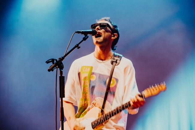 A musician from the band Wunderhorse is in sunglasses passionately sings and plays electric guitar on stage, wearing a graphic T-shirt. Vivid stage lights create a dynamic atmosphere.