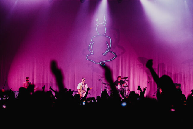The band Wunderhorse performs on stage with purple lighting and a large neon devil outline backdrop. Silhouetted fans raise hands, capturing an energetic concert vibe.