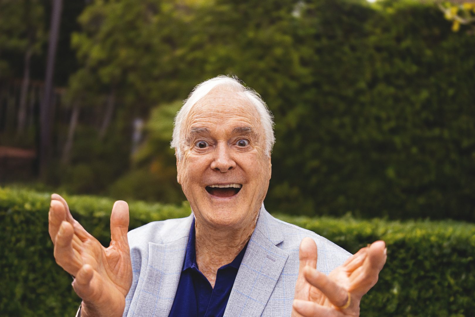 Actor and writer John Cleese in a light suit with an expressive, joyful face has his hands raised in surprise or excitement against a lush, green garden background.