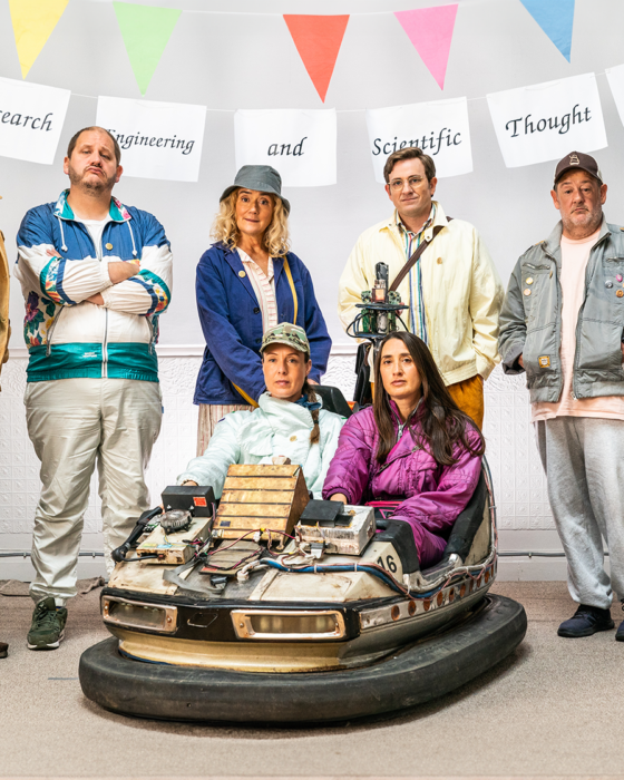 A diverse group of seven people stands in a line indoors, above them a banner reads "British Research Engineering and Scientific Thought Society." They are posing confidently, with two seated in a modified bumper car; colorful flags are hung above. The scene conveys a quirky and humorous tone.
