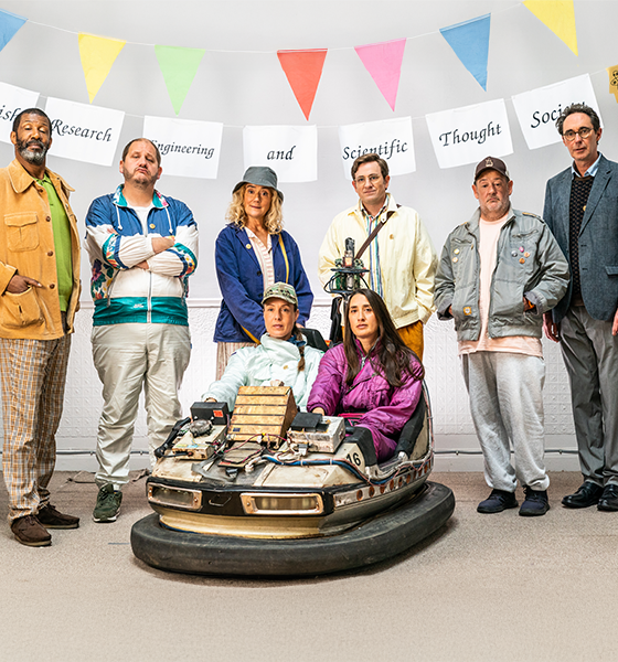 A diverse group of seven people stands in a line indoors, above them a banner reads "British Research Engineering and Scientific Thought Society." They are posing confidently, with two seated in a modified bumper car; colorful flags are hung above. The scene conveys a quirky and humorous tone.