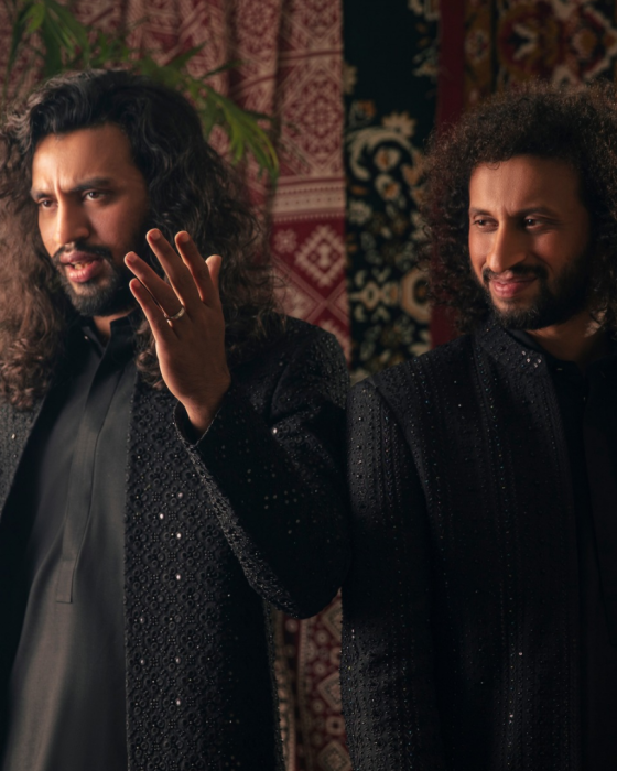Two men with long curly hair wear black, intricately detailed outfits, standing against a backdrop of rich, patterned tapestries. Warm, expressive ambiance.