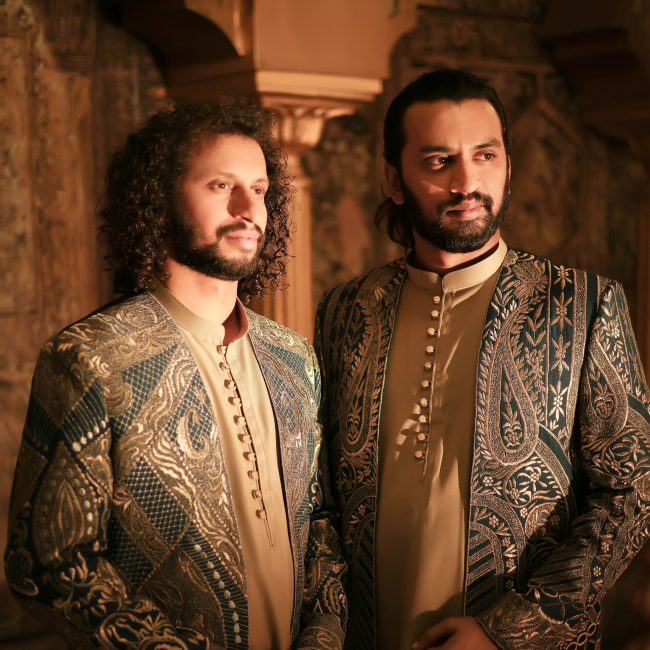 Two men in ornate, embroidered jackets stand side by side in a warmly lit room. Their composed expressions reflect elegance and confidence.