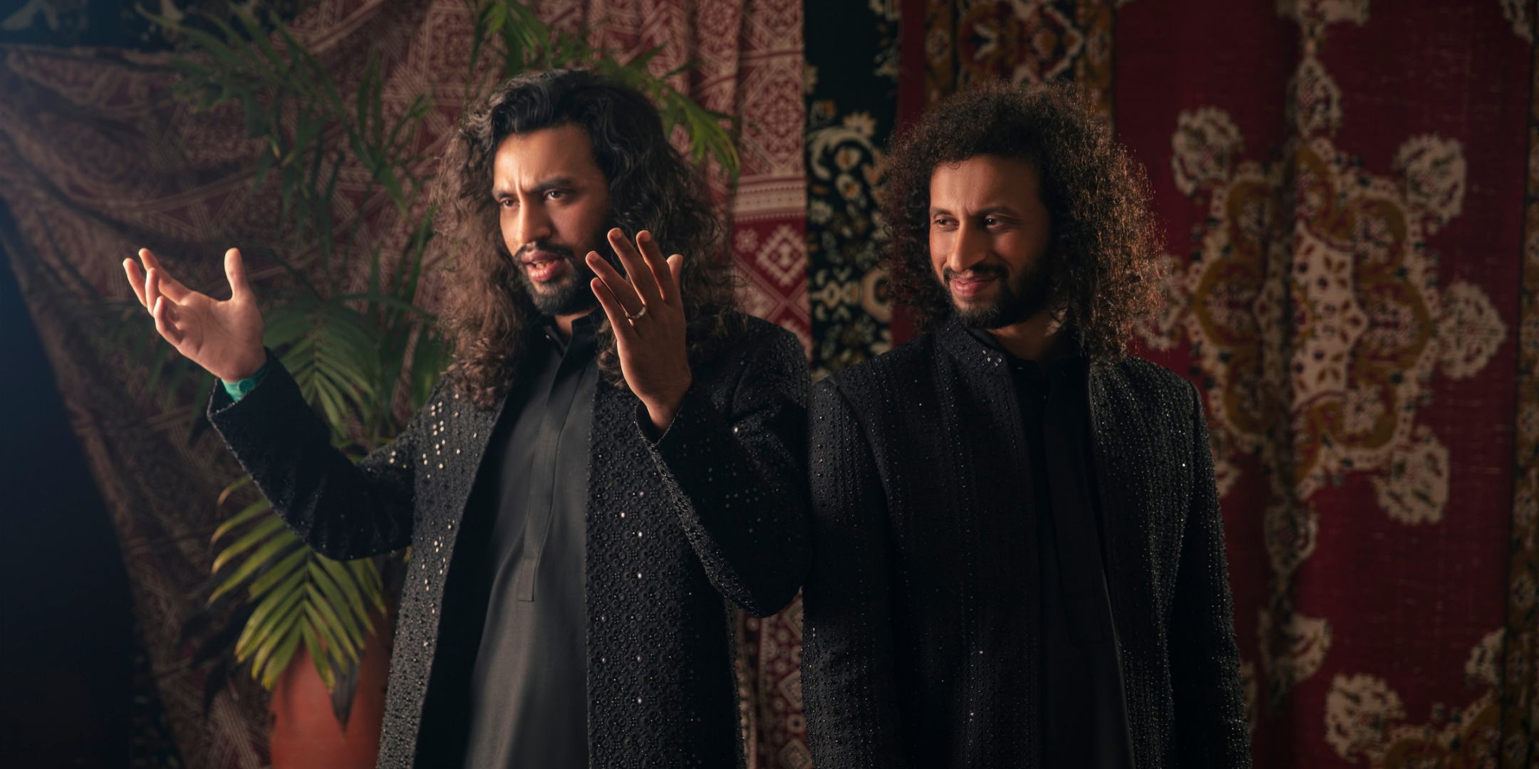 Two men with long curly hair wear black, intricately detailed outfits, standing against a backdrop of rich, patterned tapestries. Warm, expressive ambiance.