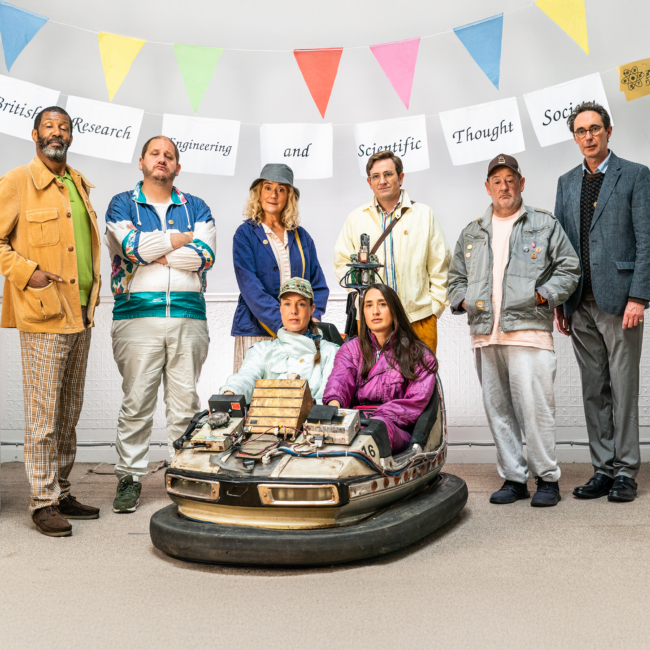 A diverse group of seven people stands in a line indoors, above them a banner reads "British Research Engineering and Scientific Thought Society." They are posing confidently, with two seated in a modified bumper car; colorful flags are hung above. The scene conveys a quirky and humorous tone.