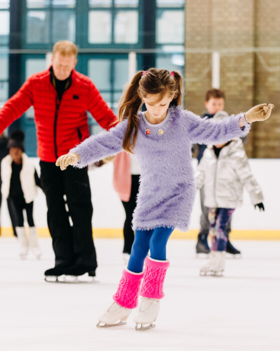 Young girl in a purple outfit ice skating, surrounded by others on the rink. She is focused, with vibrant leg warmers, creating a playful mood.