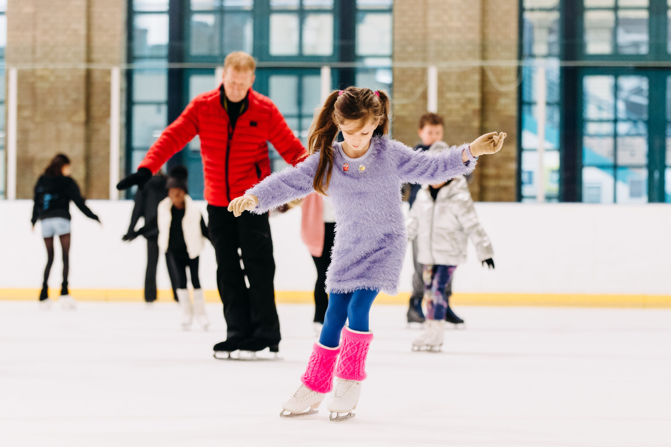 Young girl in a purple outfit ice skating, surrounded by others on the rink. She is focused, with vibrant leg warmers, creating a playful mood.