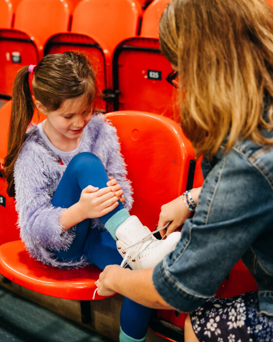 A girl in a fluffy purple sweater sits on red stadium seats, smiling as a woman in a denim jacket helps her put on white ice skates.