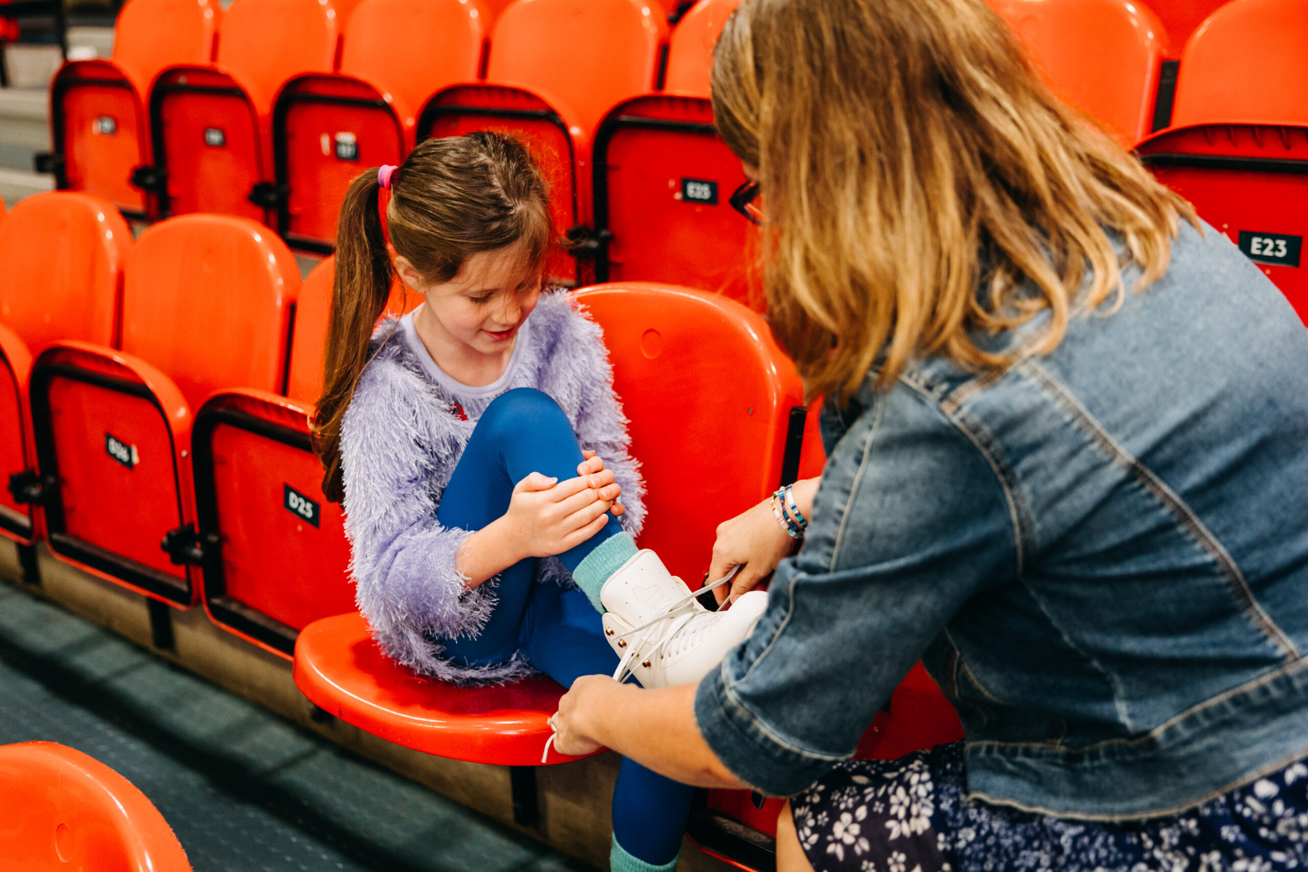A girl in a fluffy purple sweater sits on red stadium seats, smiling as a woman in a denim jacket helps her put on white ice skates.
