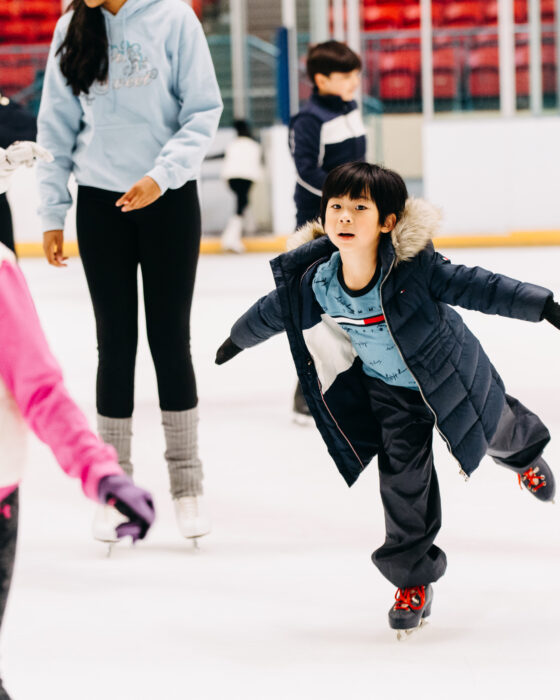 Children ice skating in an indoor rink, wearing winter jackets and gloves. The scene is lively and joyful, with a focus on a child gliding gracefully.