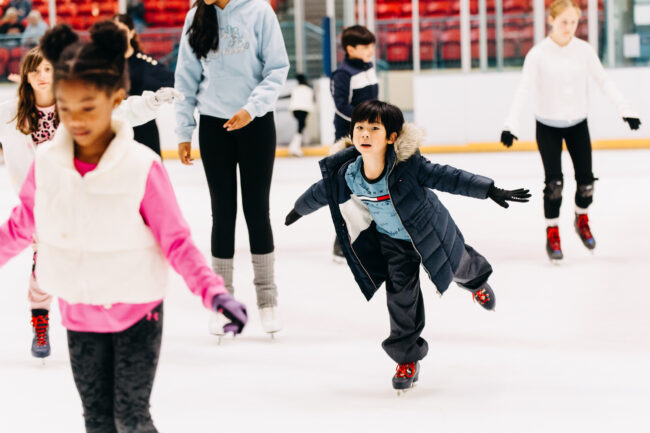 Children ice skating in an indoor rink, wearing winter jackets and gloves. The scene is lively and joyful, with a focus on a child gliding gracefully.