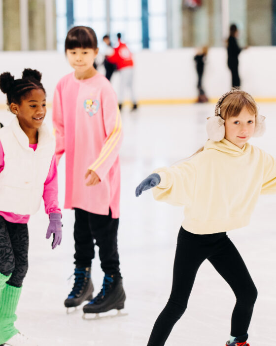 Children joyfully ice skating indoors. A girl in a yellow hoodie and earmuffs leads, while others in colorful outfits follow, all smiling and having fun.