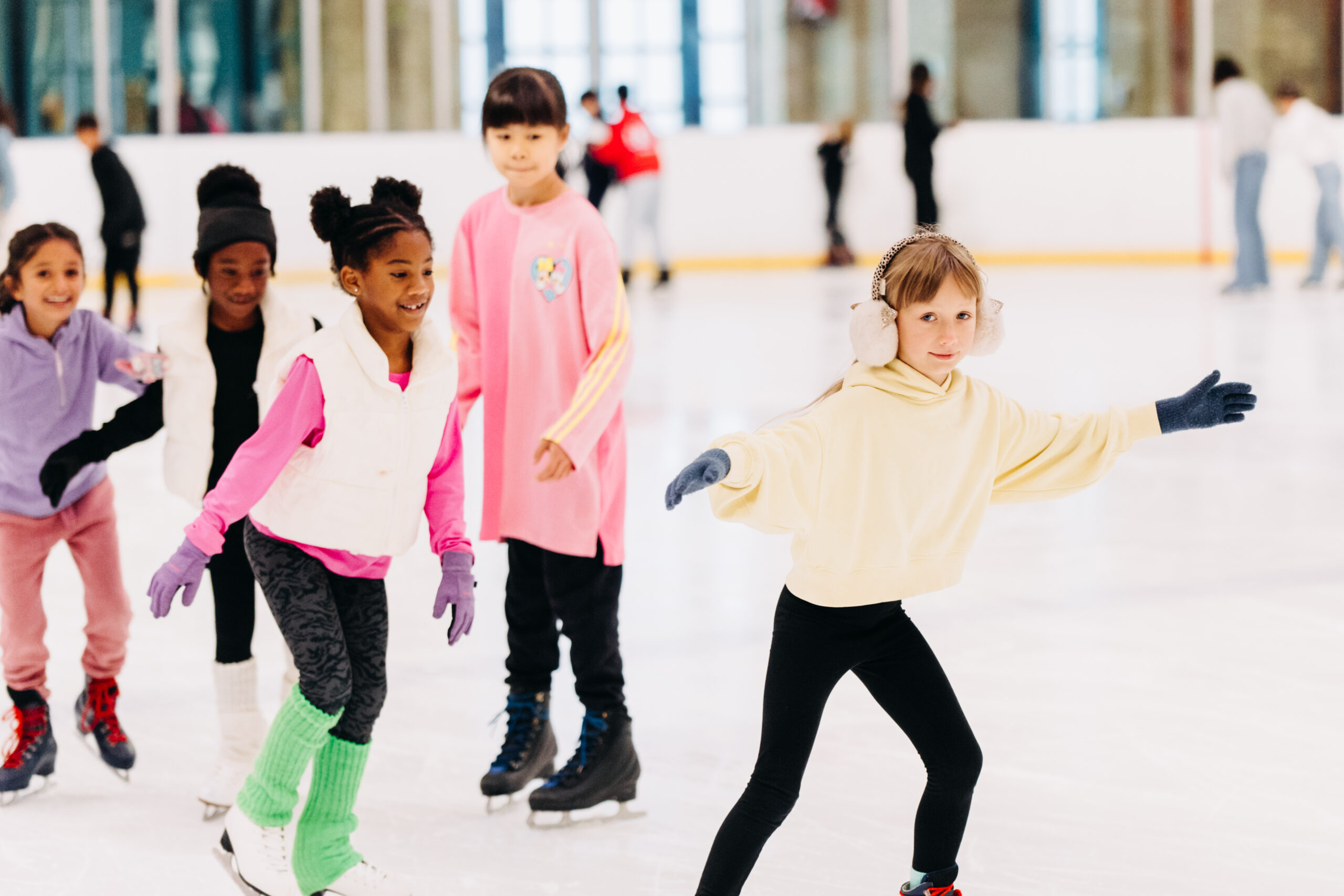 Children joyfully ice skating indoors. A girl in a yellow hoodie and earmuffs leads, while others in colorful outfits follow, all smiling and having fun.