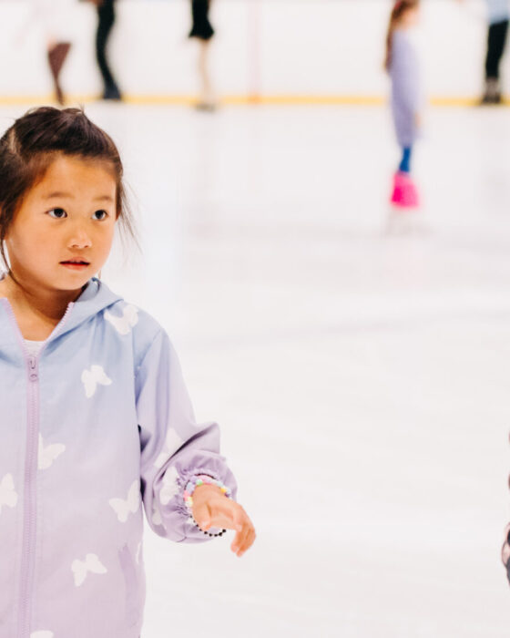 A young girl in a purple jacket and pink pants ice skates in a rink, guided by an adult. The scene conveys learning and excitement.