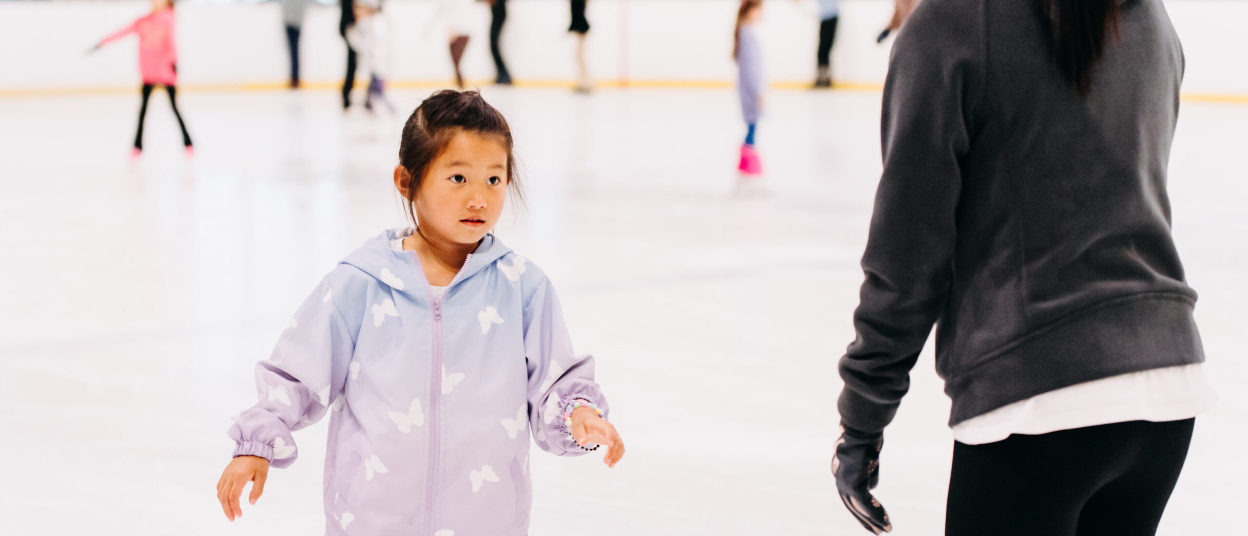 A young girl in a purple jacket and pink pants ice skates in a rink, guided by an adult. The scene conveys learning and excitement.