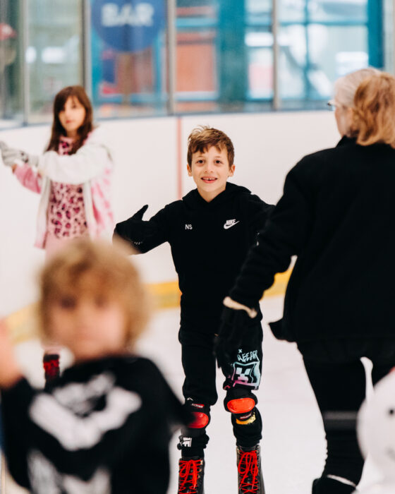A group of children ice skating indoors. A boy in a black outfit, smiling, skates confidently while others, including a girl in a pink jacket, practice nearby.