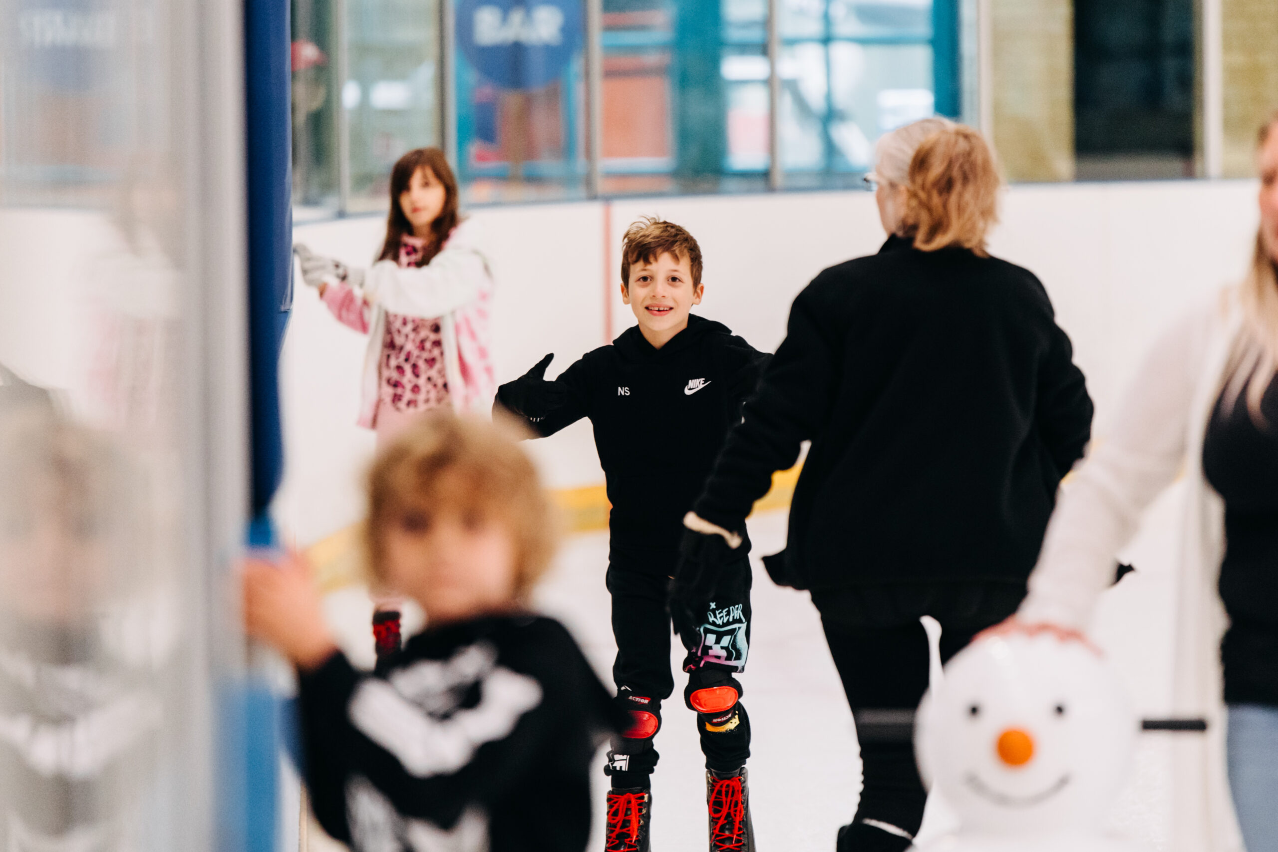 A group of children ice skating indoors. A boy in a black outfit, smiling, skates confidently while others, including a girl in a pink jacket, practice nearby.