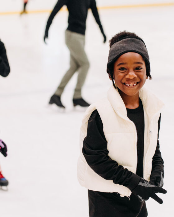 Children enjoy ice skating indoors. A smiling girl in a white vest is foregrounded, with two other kids skating behind her. The scene is cheerful and lively.