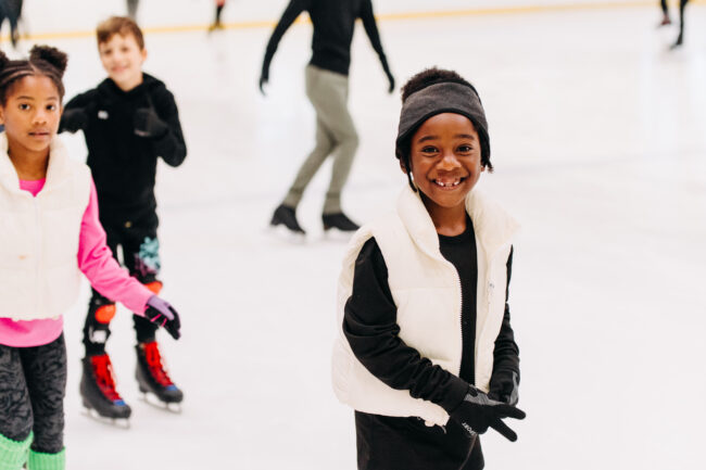 Children enjoy ice skating indoors. A smiling girl in a white vest is foregrounded, with two other kids skating behind her. The scene is cheerful and lively.