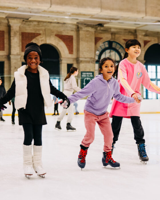 Five children in colourful sweaters and ice skates joyfully hold hands while gliding on an indoor ice rink, with soft natural light filtering in.