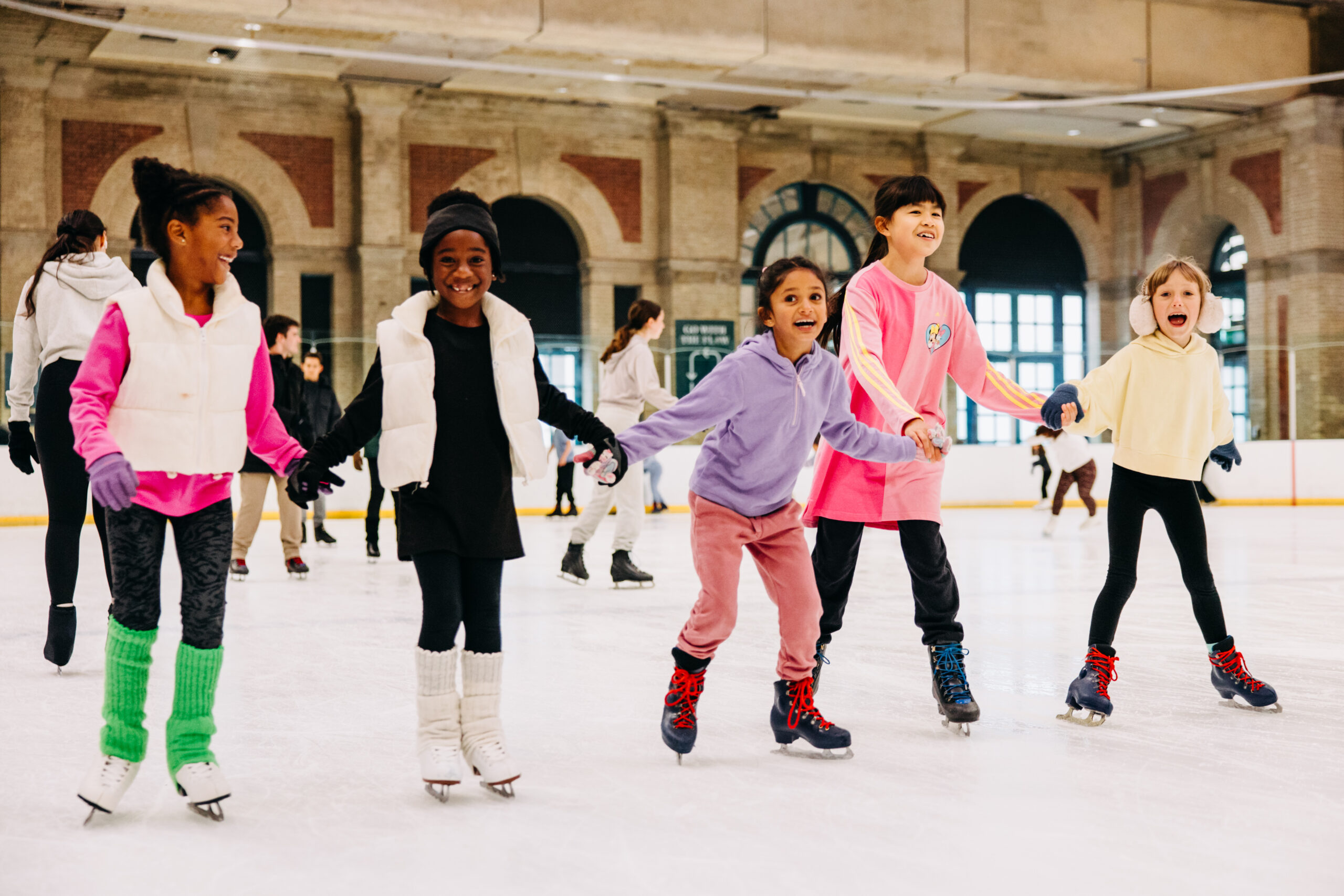 Five children in colourful sweaters and ice skates joyfully hold hands while gliding on an indoor ice rink, with soft natural light filtering in.