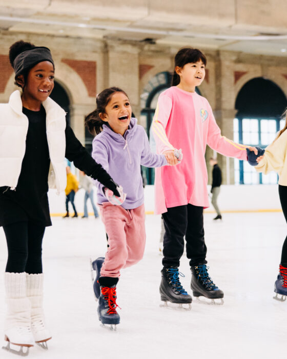 Four children ice skating indoors, holding hands, and smiling. They wear colourful winter clothes, and the backdrop features large windows and brick arches.