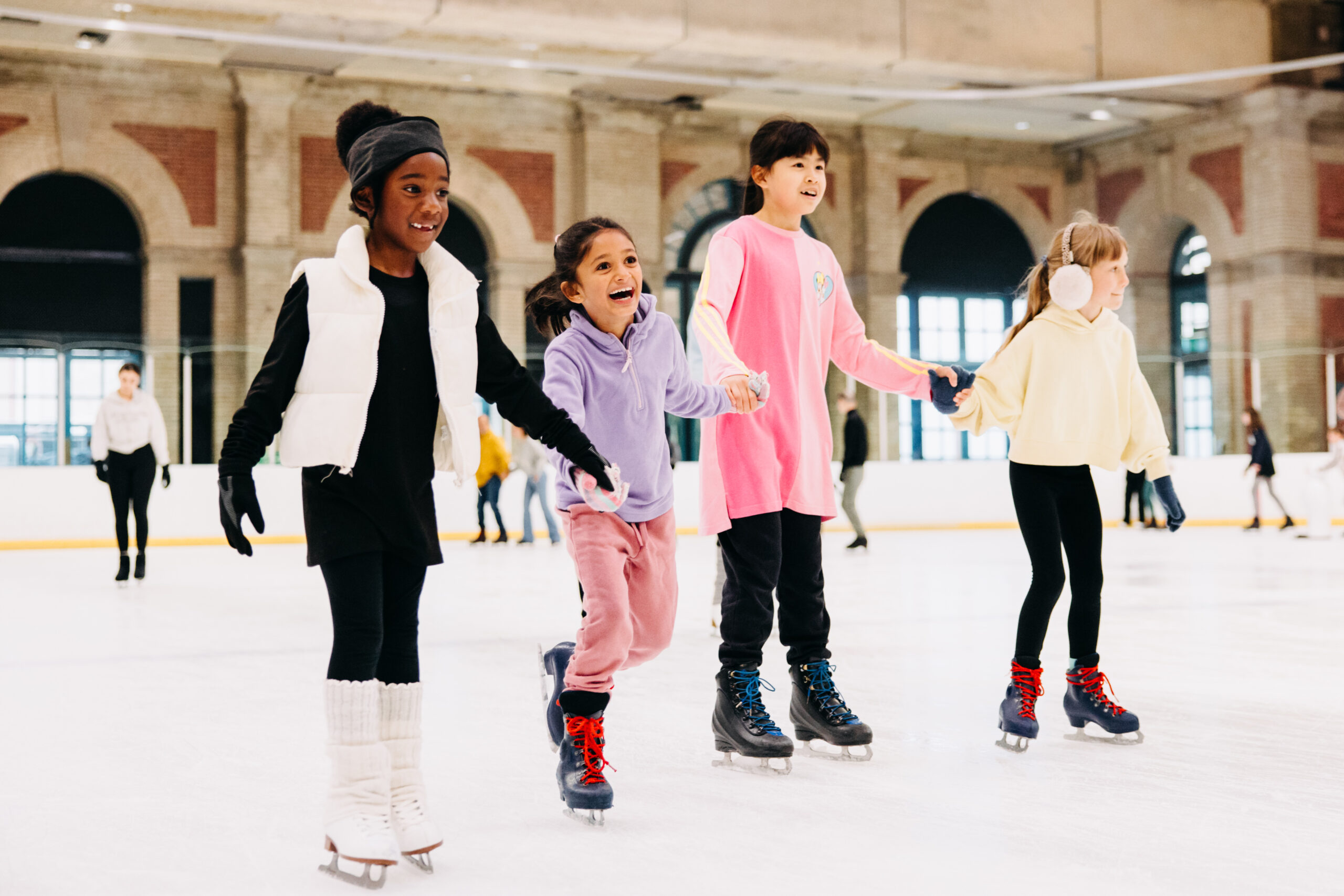 Four children ice skating indoors, holding hands, and smiling. They wear colourful winter clothes, and the backdrop features large windows and brick arches.